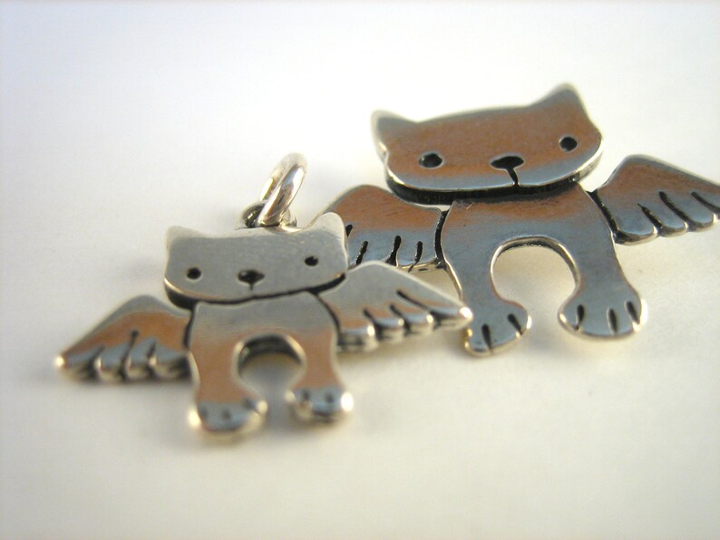 Puede incluir: Dos encantos de plata en forma de gatos con alas. El encanto m&aacute;s grande es ligeramente m&aacute;s grande que el encanto m&aacute;s peque&ntilde;o.