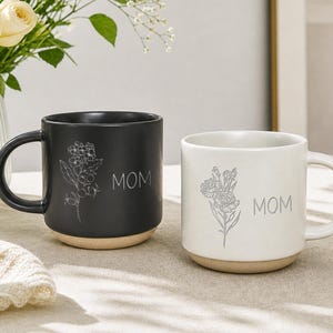 Könnte beinhalten: Ein mattschwarzer Keramikbecher und ein glänzend weißer Keramikbecher, beide mit einer feinen floralen Strichzeichnung und dem Wort MOM in einer einfachen serifenlosen Schrift. Beide Becher haben einen naturbelassenen, unglasierten Boden.
