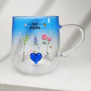 May include: A clear glass mug with a blue gradient, featuring the words "First Mom" and floral illustrations. The mug has a handle and is decorated with butterflies and the names "Marian" and "Eliza".