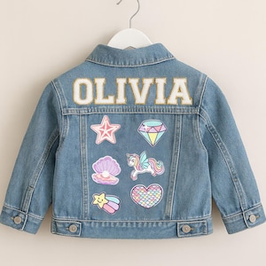 Puede incluir: Chaqueta vaquera azul claro con el nombre "OLIVIA" en letras blancas y doradas en la espalda. La chaqueta presenta parches coloridos de una estrella, un diamante, una concha, un unicornio, una estrella fugaz y un corazón. La chaqueta está colgada en una percha blanca.