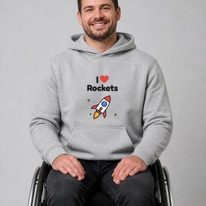 May include: A light gray hooded sweatshirt featuring a graphic print on the chest that displays the text I, a red heart symbol, the word Rockets, and a colorful illustration of a rocket ship surrounded by small four-pointed star icons.