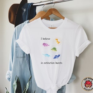 Peut inclure: T-shirt blanc avec un motif de dinosaures colorés et le texte "Je crois aux explosions d'extinction".