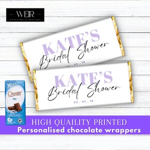 May include: Two white chocolate bar wrappers with gold foil accents. The wrappers are personalised with the text "KATE'S Bridal Shower 05.05.18" in purple and black lettering.