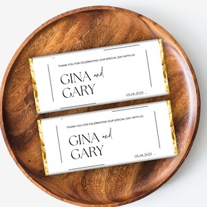 May include: Two white chocolate bar wrappers with gold foil accents. The wrappers have black text that reads "Thank you for celebrating our special day with us."  "GINA and GARY" and "05.08.2023".