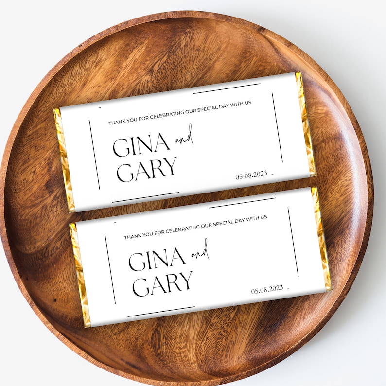 May include: Two white chocolate bar wrappers with gold foil accents. The wrappers have black text that reads "Thank you for celebrating our special day with us."  "GINA and GARY" and "05.08.2023".