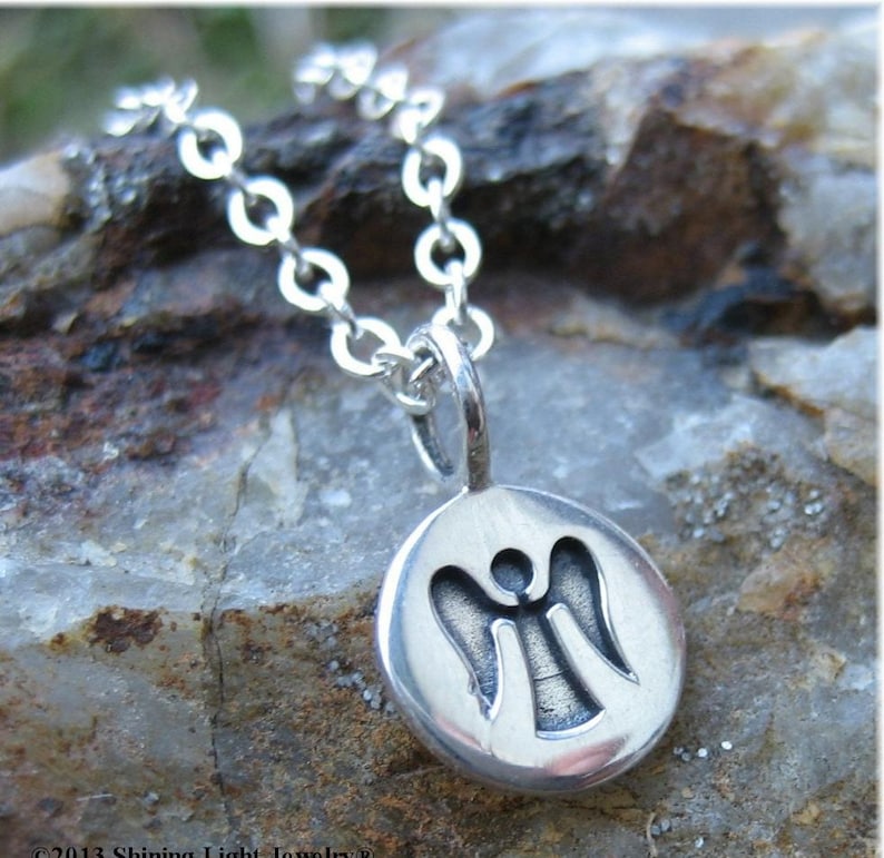 Pu&ograve; includere: Una collana d'argento con un ciondolo rotondo raffigurante un angelo. Il ciondolo &egrave; attaccato a una catena delicata. La collana &egrave; esposta su una superficie di pietra strutturata grigia e marrone.