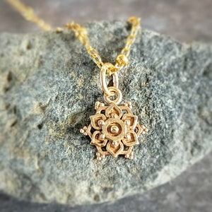 A small gold colored lotus flower mandala charm hangs on a gold chain against a grey rock backdrop.