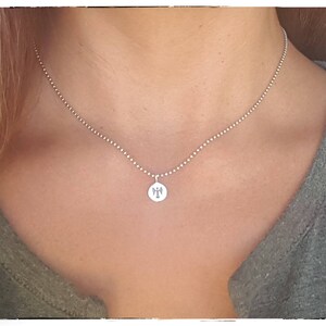 Pu&ograve; includere: Una collana color argento con un piccolo pendente rotondo. Il pendente presenta un design dettagliato. La collana &egrave; indossata sulla pelle di una persona e una maglietta grigia.