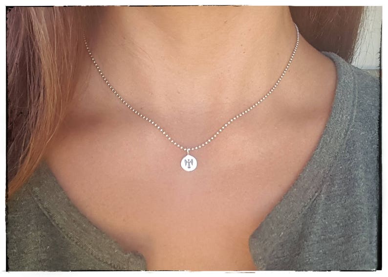 Pu&ograve; includere: Una collana color argento con un piccolo pendente rotondo. Il pendente presenta un design dettagliato. La collana &egrave; indossata sulla pelle di una persona e una maglietta grigia.