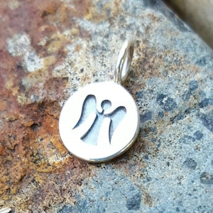 Pu&ograve; includere: Un ciondolo color argento con un design circolare con una silhouette di angelo ritagliata. Il ciondolo ha un piccolo anello nella parte superiore per una catena. Lo sfondo &egrave; una superficie di pietra strutturata, grigio-marrone.
