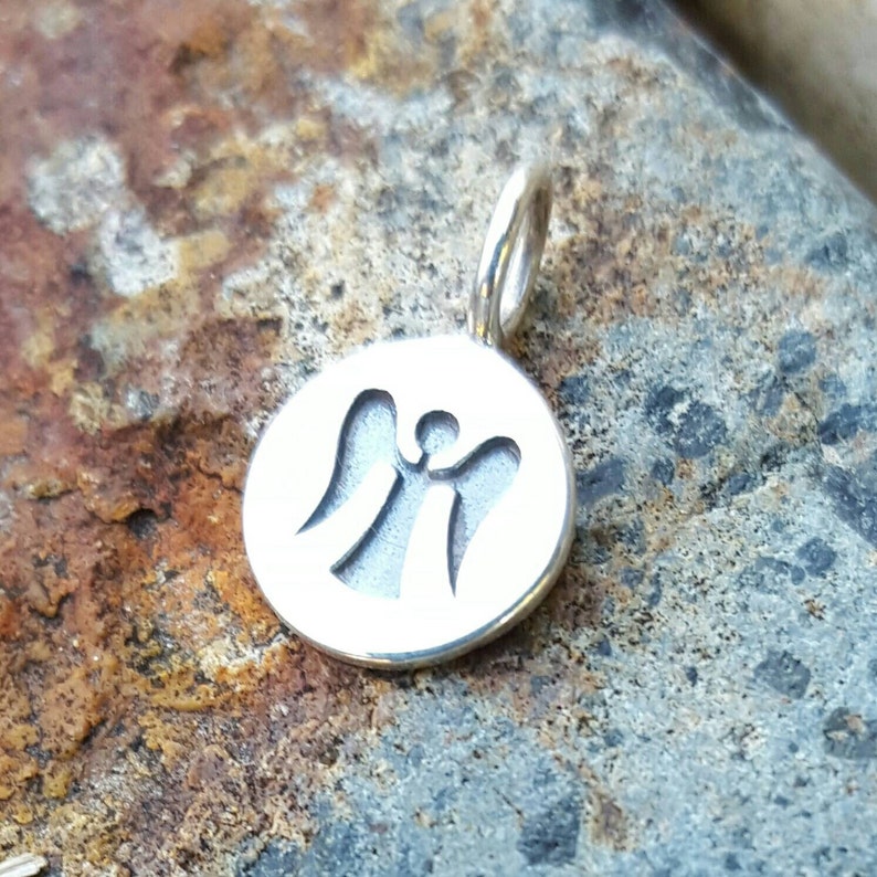 Pu&ograve; includere: Un ciondolo color argento con un design circolare con una silhouette di angelo ritagliata. Il ciondolo ha un piccolo anello nella parte superiore per una catena. Lo sfondo &egrave; una superficie di pietra strutturata, grigio-marrone.