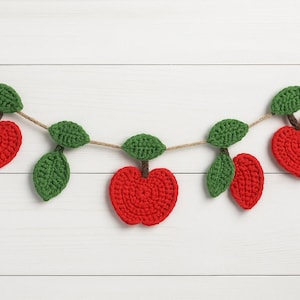 Puede incluir: Una guirnalda de ganchillo hecha a mano con formas de manzana rojas, hojas y tallos verdes. Las manzanas están ensartadas en una cuerda natural, creando un acento decorativo. Perfecto para la decoración de temporada.