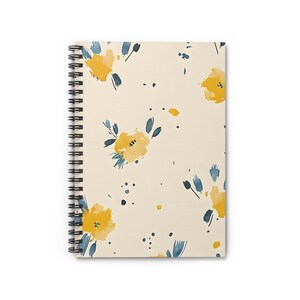 Puede incluir: Cuaderno de espiral con una cubierta de color crema con un patrón floral de acuarela. El diseño incluye flores amarillas y hojas y acentos azules. La encuadernación en espiral del cuaderno es negra.