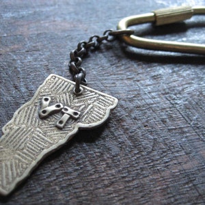 Könnte beinhalten: Ein Schlüsselanhänger aus Messing mit einer detaillierten Umrandung des Bundesstaates Vermont mit den Buchstaben "VT" in der Mitte. Der Schlüsselanhänger ist an einem Messingring mit einer Kette befestigt.