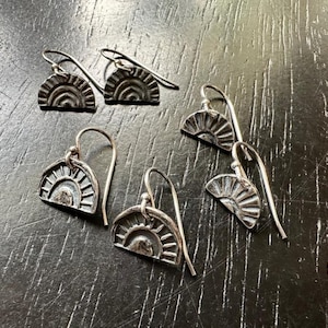 Puede incluir: Cinco pares de pendientes de plata con un diseño de medio círculo que presenta un sol con rayos. Los pendientes están sobre una superficie de madera oscura.