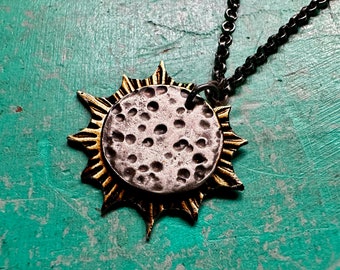 May include: A necklace featuring a circular pendant with a textured, cratered silver-toned center and a radiating gold-toned sunburst frame with sharp points, suspended from a dark, oxidized metal chain with round links.