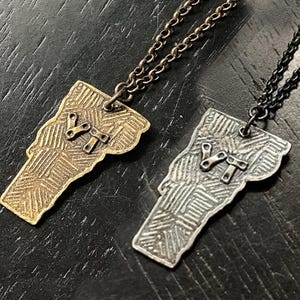 Puede incluir: Dos collares con forma del estado de Vermont, uno con una cadena dorada y un colgante de latón, el otro con una cadena negra y un colgante de plata. Ambos colgantes presentan una superficie texturizada y las letras "VT" en el centro.