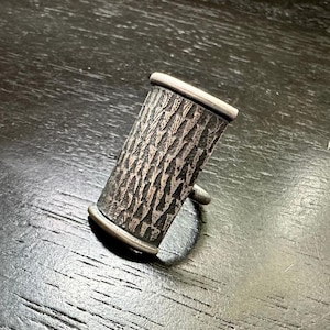 以下が含まれることがあります： 木目のテクスチャが施されたシルバーリング。リングは金属製で、シンプルなバンドです。