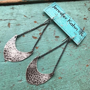 May include: A pair of silver earrings with a hammered crescent moon design, hanging from black chains. The earrings are on a turquoise blue background with a white card that says "Jennifer Kahn Jewelry".
