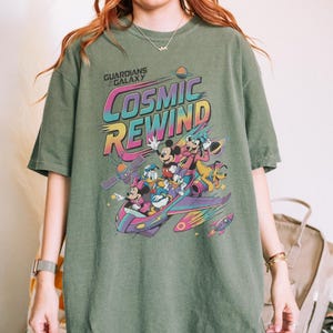 Puede incluir: Una camiseta verde oliva con un estampado gr&aacute;fico que incluye el texto Guardians of the Galaxy Cosmic Rewind. El dise&ntilde;o muestra a Mickey Mouse, Minnie Mouse, Donald Duck, Daisy Duck, Goofy y Pluto viajando en una nave espacial colorida.