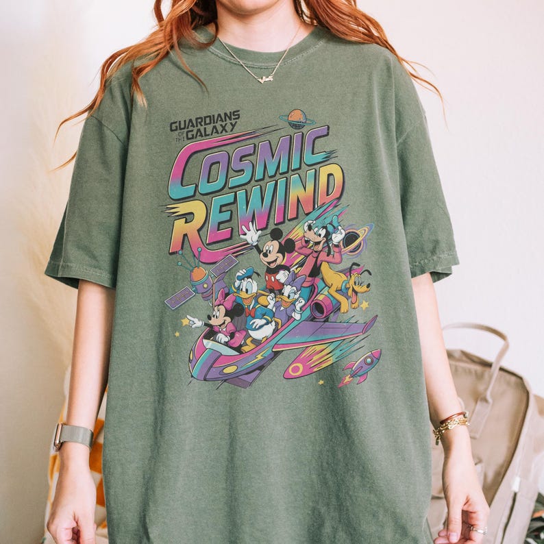 Puede incluir: Una camiseta verde oliva con un estampado gr&aacute;fico que incluye el texto Guardians of the Galaxy Cosmic Rewind. El dise&ntilde;o muestra a Mickey Mouse, Minnie Mouse, Donald Duck, Daisy Duck, Goofy y Pluto viajando en una nave espacial colorida.