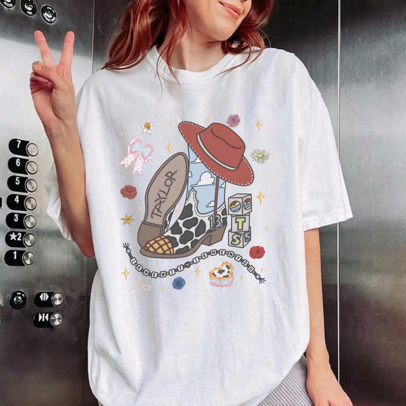 May include: A white t-shirt featuring a graphic print of a cowboy boot with a cow print pattern, a red cowboy hat, a pink bow, and stacked blocks with the letters T, S, and the number 13. The word TAYLOR is printed on the boot, and a beaded bracelet graphic surrounds the bottom.