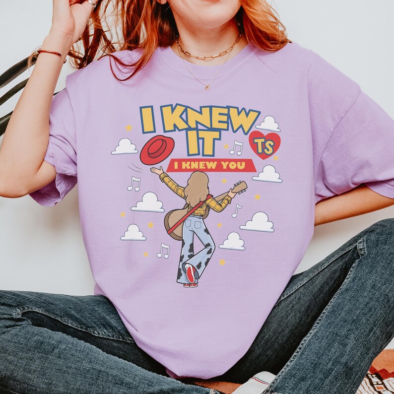 K&ouml;nnte beinhalten: Ein hellviolettes T-Shirt mit einer Grafik einer langhaarigen Person, die Gitarre spielt und ein gelbes Karohemd sowie eine Schlaghose mit Kuhfleckenmuster tr&auml;gt. Der Text lautet I KNEW IT I KNEW YOU, erg&auml;nzt durch einen roten Hut und Wolken.