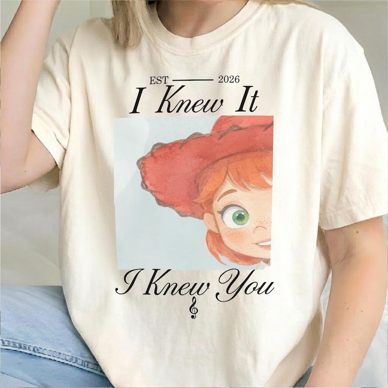May include: A cream-colored short-sleeve t-shirt featuring a graphic print of a cartoon character with red hair and a red hat. The text reads EST 2026 I Knew It I Knew You with a small musical treble clef symbol below the bottom line of text.