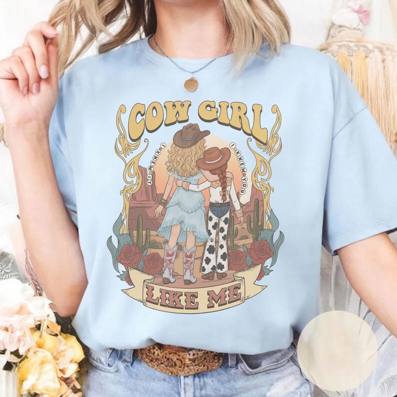 May include: A light blue t-shirt featuring a graphic print of two figures in cowboy hats standing in a desert landscape with cacti and roses. The text reads COW GIRL and LIKE ME, with the phrase I KNEW YOU curved around the central illustration.