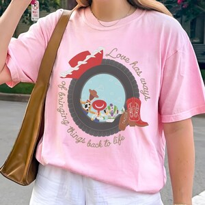 May include: A light pink t-shirt featuring a graphic of a dark gray tire framing a scene with cartoon characters. The design includes a red cowboy hat, a red cowboy boot with the text Taylor, and the phrase Love has ways of bringing things back to life.