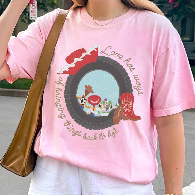 May include: A light pink t-shirt featuring a graphic of a dark gray tire framing a scene with cartoon characters. The design includes a red cowboy hat, a red cowboy boot with the text Taylor, and the phrase Love has ways of bringing things back to life.
