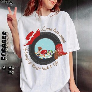 May include: A white t-shirt featuring a graphic of a black tire framing a light blue sky scene with cartoon characters. The design includes a red cowboy hat, a red cowboy boot with the name Taylor, and the text Love has ways of bringing things back to life.