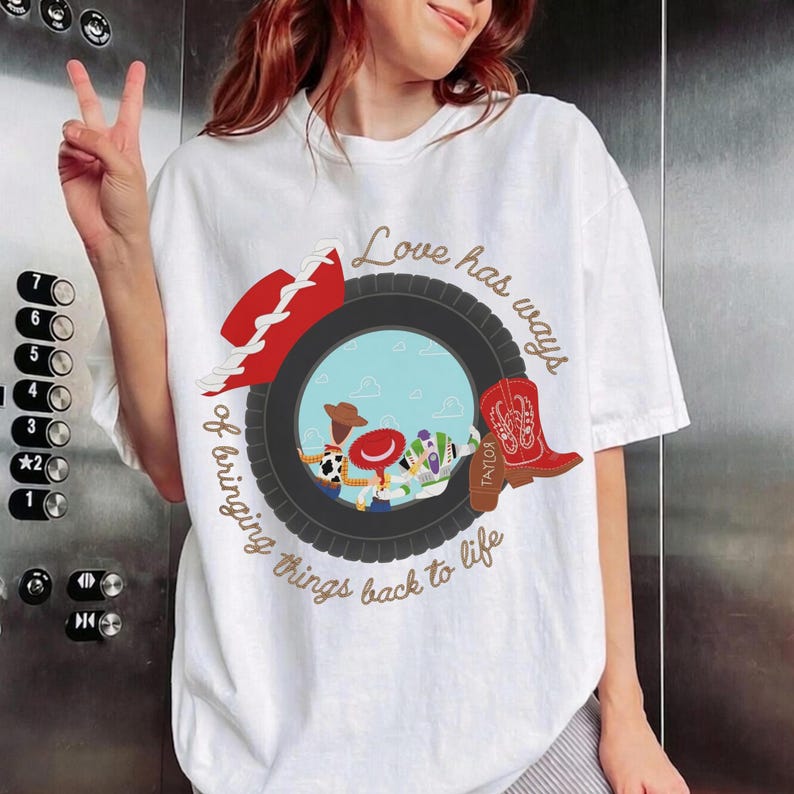 May include: A white t-shirt featuring a graphic of a black tire framing a light blue sky scene with cartoon characters. The design includes a red cowboy hat, a red cowboy boot with the name Taylor, and the text Love has ways of bringing things back to life.