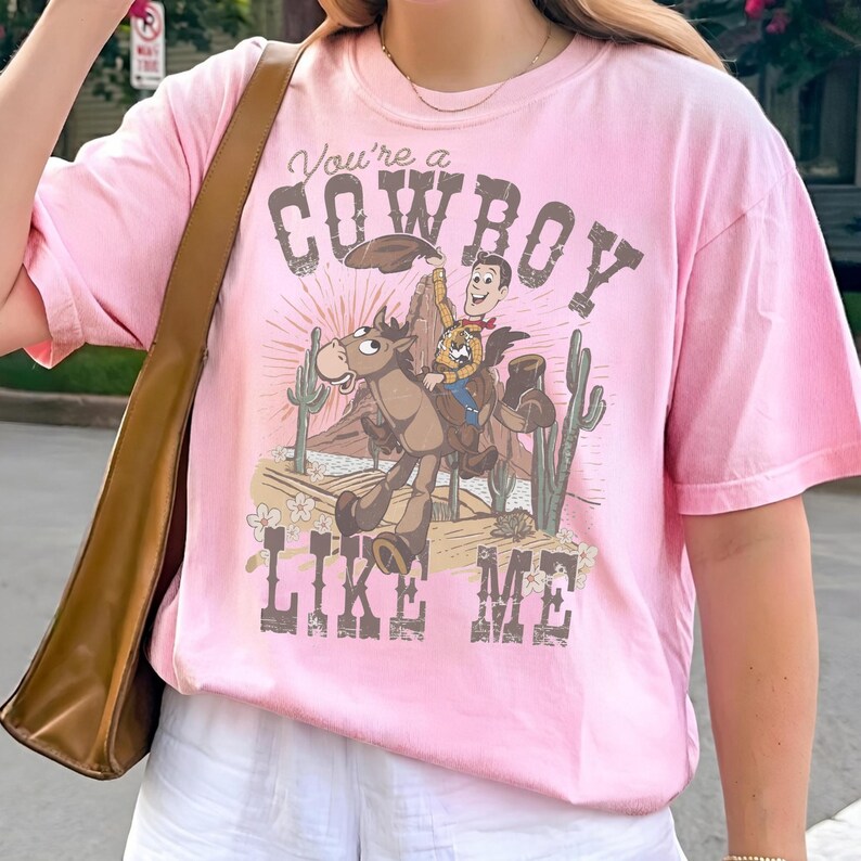 Puede incluir: Una camiseta rosa claro de manga corta con un estampado gr&aacute;fico de un vaquero de dibujos animados montando a caballo en un paisaje des&eacute;rtico con cactus. El texto "You're a COWBOY LIKE ME" est&aacute; impreso en una fuente marr&oacute;n desgastada.
