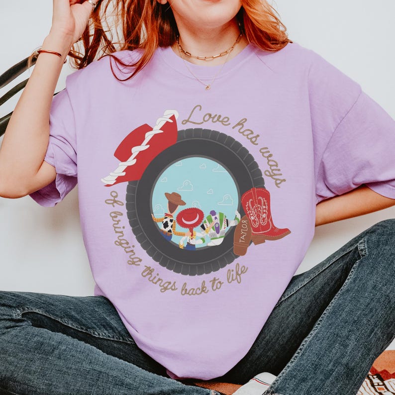 May include: A light purple t-shirt featuring a graphic of a black tire surrounding a scene with three cartoon characters. A red cowboy hat and a red cowboy boot are placed on the tire. The text reads, Love has ways of bringing things back to life.