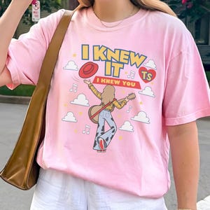 K&ouml;nnte beinhalten: Ein hellrosa T-Shirt mit einer grafischen Illustration einer Person mit langem Haar, die eine Akustikgitarre spielt und ein gelbes Karohemd sowie eine Hose mit Kuhfleckenmuster tr&auml;gt. Der Text lautet I KNEW IT I KNEW YOU TS.