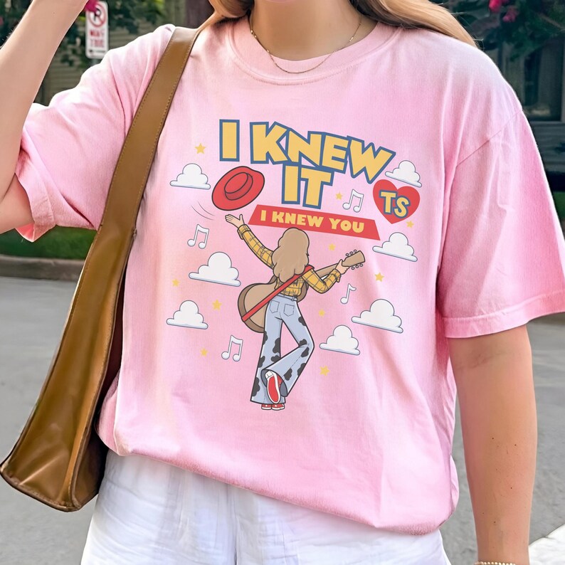 K&ouml;nnte beinhalten: Ein hellrosa T-Shirt mit einer grafischen Illustration einer Person mit langem Haar, die eine Akustikgitarre spielt und ein gelbes Karohemd sowie eine Hose mit Kuhfleckenmuster tr&auml;gt. Der Text lautet I KNEW IT I KNEW YOU TS.