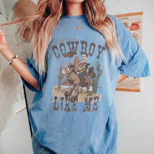 Puede incluir: Una camiseta de manga corta de color azul claro con un estampado gr&aacute;fico de un vaquero montando a caballo en un paisaje des&eacute;rtico con cactus. El texto "You're a COWBOY LIKE ME" est&aacute; impreso en letras marrones sobre la ilustraci&oacute;n.