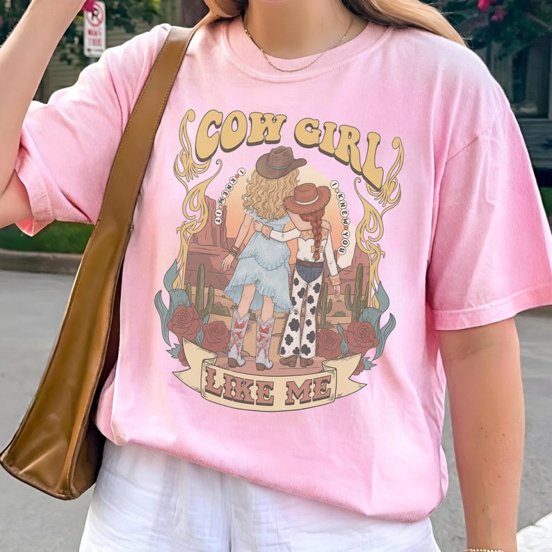 May include: A light pink short-sleeve t-shirt featuring a graphic print of two figures in cowboy hats standing in a desert landscape with cacti and roses. The text reads COW GIRL and LIKE ME, with I KNEW YOU written in small vertical arcs.