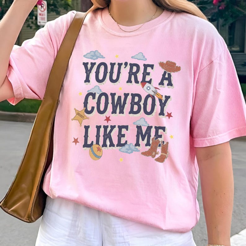 May include: A light pink short-sleeve t-shirt featuring the dark blue text "YOU'RE A COWBOY LIKE ME" surrounded by graphic illustrations of a brown cowboy hat, a rocket ship, a yellow sheriff star, a colorful ball, small clouds, and red stars.