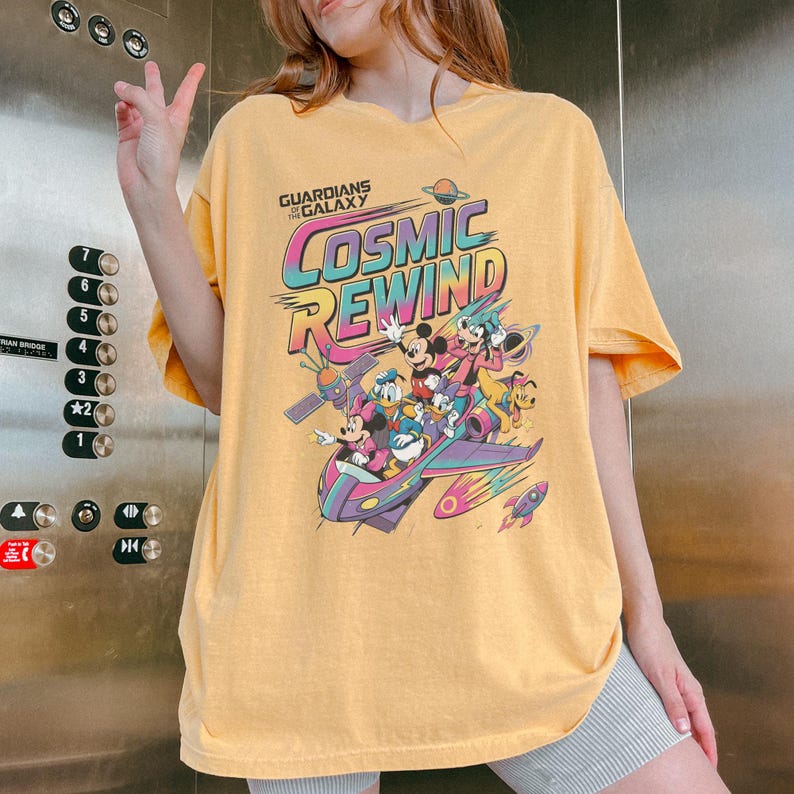 Puede incluir: Una camiseta amarilla de manga corta con un estampado gr&aacute;fico que incluye el texto Guardians of the Galaxy Cosmic Rewind. El dise&ntilde;o muestra ilustraciones coloridas de Mickey Mouse, Minnie Mouse, Donald Duck, Daisy Duck, Goofy y Pluto en una nave.