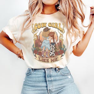 May include: A cream-colored short-sleeve t-shirt featuring a graphic print of two figures in cowboy hats and boots standing in a desert landscape with cacti and roses. The text COW GIRL LIKE ME is printed in large, stylized gold-toned lettering.