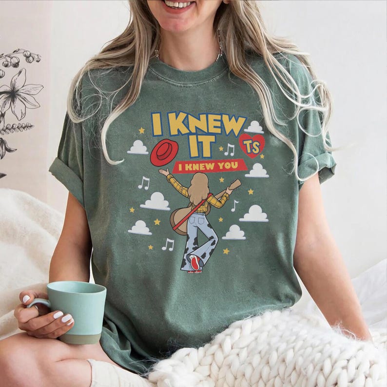 K&ouml;nnte beinhalten: Ein salbeigr&uuml;nes T-Shirt mit einer Grafik einer Person mit langem Haar, die eine Akustikgitarre spielt. Das Design enth&auml;lt den Text I KNEW IT und I KNEW YOU, einen roten Hut, ein rotes Herz, Musiknoten, Sterne und wei&szlig;e Wolken auf gr&uuml;nem Grund.