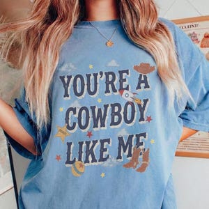 May include: A blue short-sleeve t-shirt featuring the text "YOU'RE A COWBOY LIKE ME" in dark blue letters. The design includes a brown cowboy hat, a rocket ship, a yellow sheriff star, a ball, and brown cowboy boots with small stars and clouds.