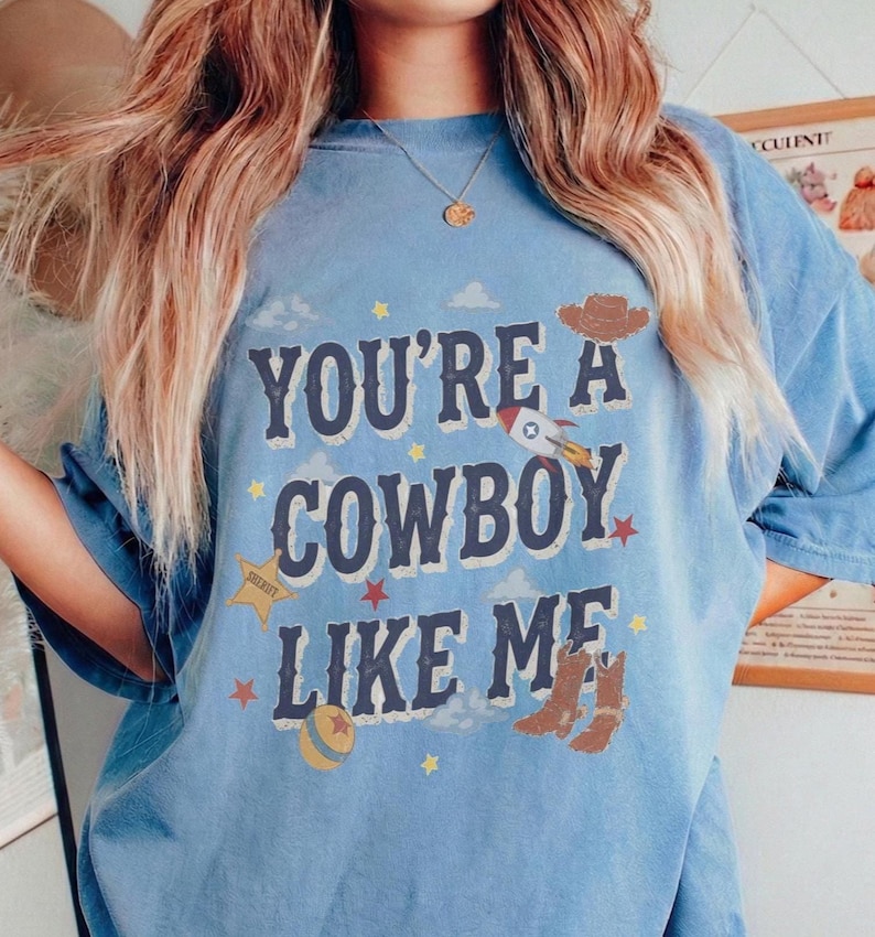 May include: A blue short-sleeve t-shirt featuring the text "YOU'RE A COWBOY LIKE ME" in dark blue letters. The design includes a brown cowboy hat, a rocket ship, a yellow sheriff star, a ball, and brown cowboy boots with small stars and clouds.