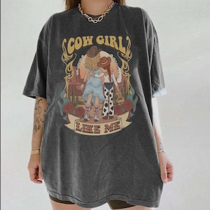 May include: A charcoal gray short-sleeve t-shirt featuring a graphic print of two figures in cowboy hats embracing in a desert landscape with cacti and roses. The text COW GIRL and LIKE ME is printed in a yellow font above and below the illustration.