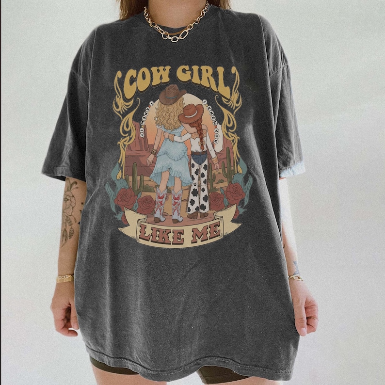 May include: A charcoal gray short-sleeve t-shirt featuring a graphic print of two figures in cowboy hats embracing in a desert landscape with cacti and roses. The text COW GIRL and LIKE ME is printed in a yellow font above and below the illustration.