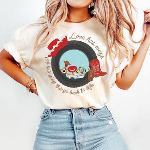 May include: A cream-colored short-sleeve t-shirt featuring a graphic of a black tire framing cartoon characters. The design includes a red hat, a red cowboy boot with the name Taylor, and the text Love has ways of bringing things back to life.