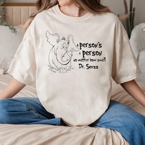 May include: Cream-colored t-shirt featuring a black and white illustration of an elephant-like creature with large ears. The shirt includes the quote "A person's a person no matter how small" by Dr. Seuss.