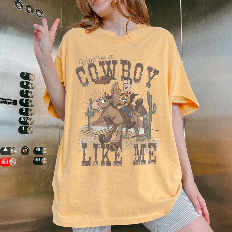 Puede incluir: Una camiseta de manga corta color amarillo mostaza con un estampado gr&aacute;fico del personaje Woody montando un caballo en un paisaje des&eacute;rtico con cactus. El texto "You're a COWBOY LIKE ME" est&aacute; impreso en una fuente marr&oacute;n oscuro desgastada.